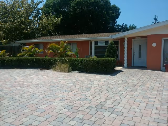 Front exterior view of a single-story building with a paved driveway, trimmed hedges, palm plants, and trees in the background under a clear blue sky.
