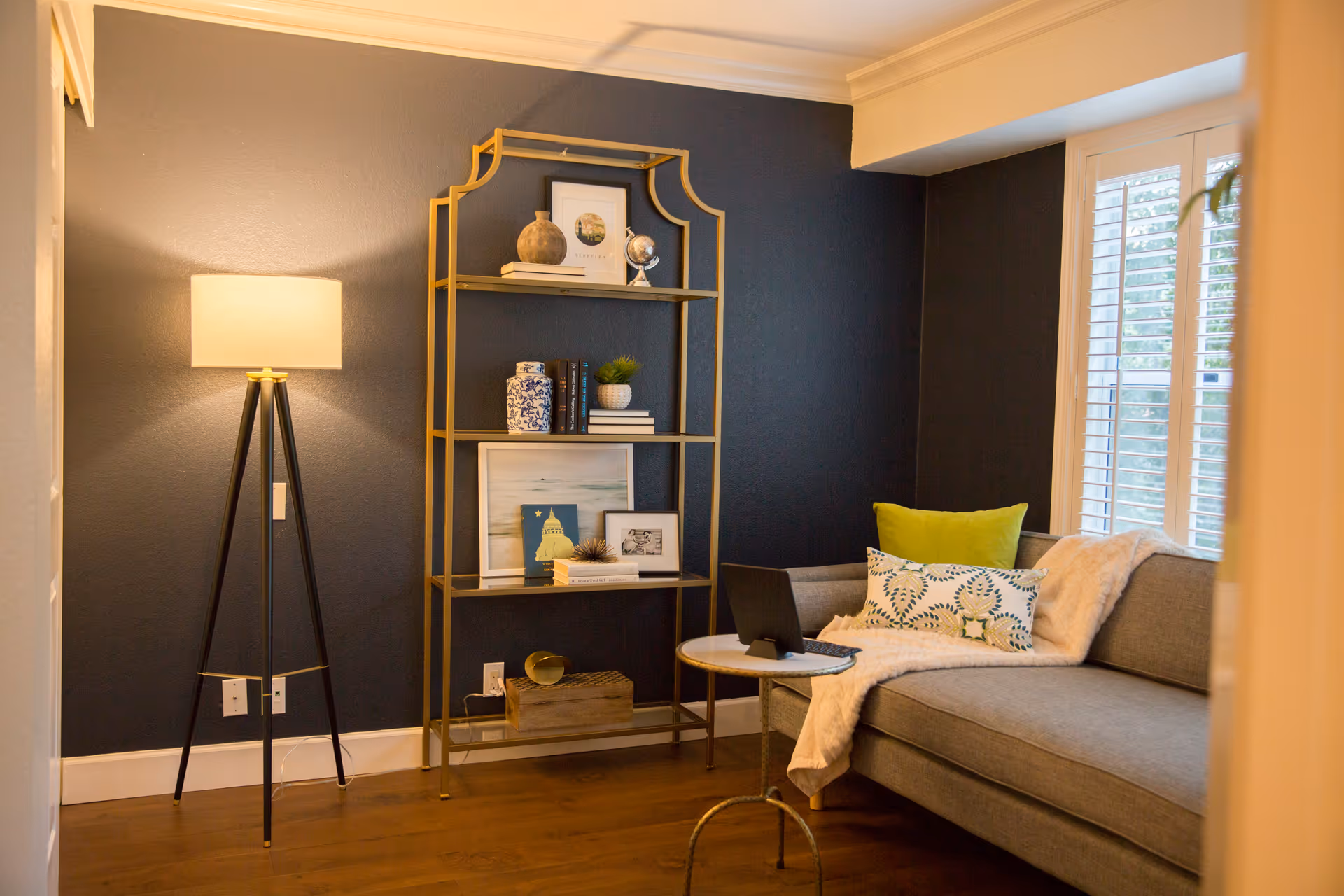 A cozy living room corner with a gray sofa adorned with green and patterned pillows and a white throw blanket. A small round table in front of the sofa holds a laptop. To the left, a tall black tripod floor lamp with a beige lampshade stands next to a gold metal shelving unit displaying decorative items and framed pictures. The walls are painted dark blue, and a window with white shutters lets in natural light.