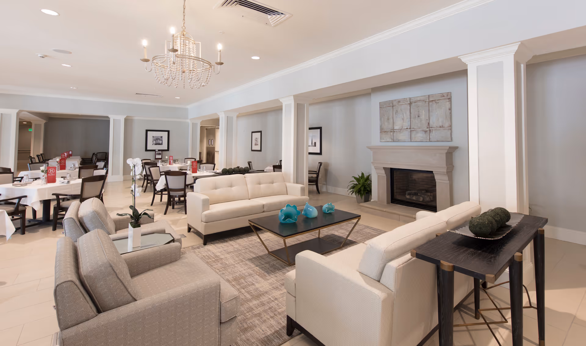 Bright communal living room with sofas, armchairs, a coffee table, fireplace, and dining tables in the background.