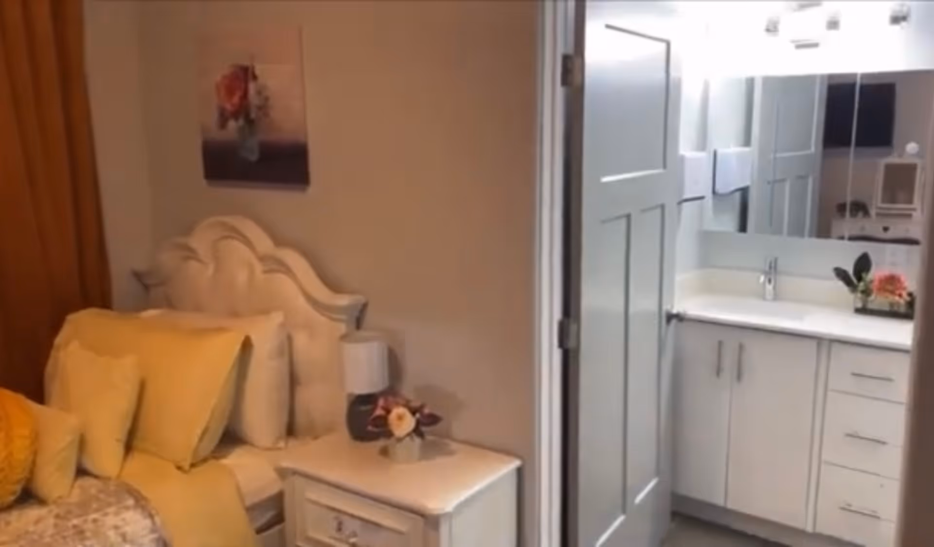 Small bedroom with a decorative headboard, bedside table and lamp next to an open door revealing a white bathroom vanity.