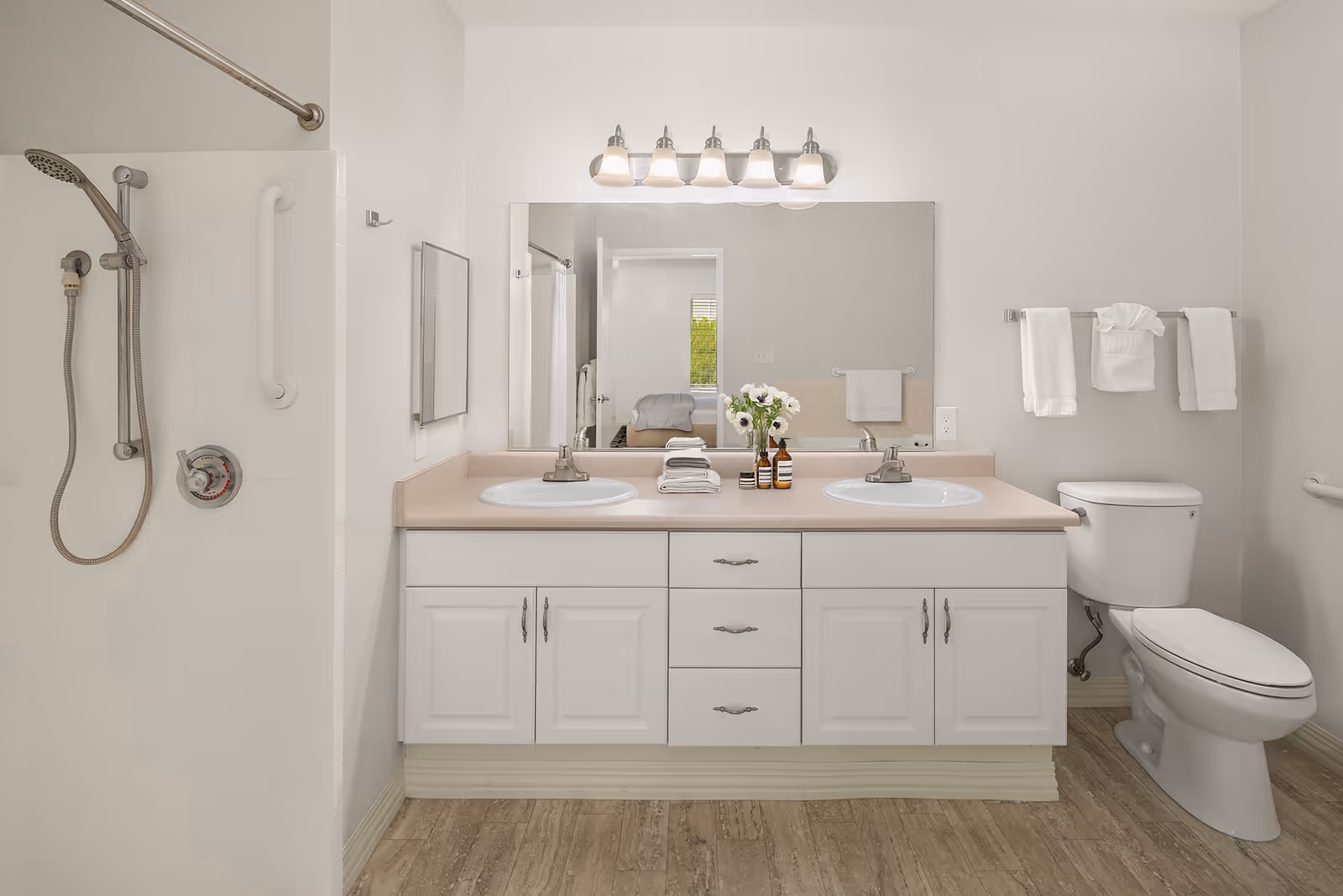 A clean and modern bathroom featuring a double sink vanity with white cabinets and a beige countertop. Above the sinks is a large mirror with a light fixture containing five bulbs. To the left is a walk-in shower with a handheld showerhead and a grab bar. To the right is a white toilet with a towel rack above it holding three neatly folded white towels. The floor has a wood-like finish.