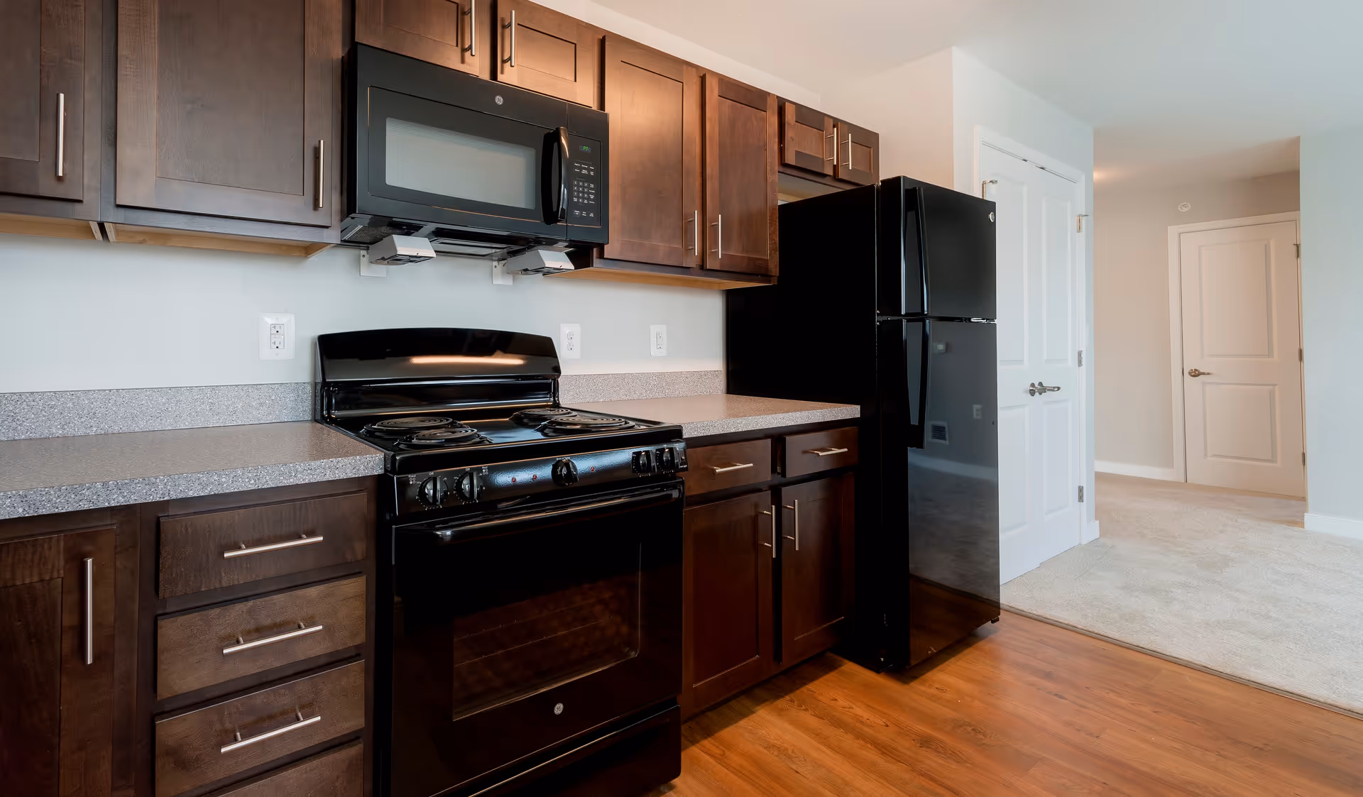 Modern kitchen with dark wood cabinets, black stove, microwave and refrigerator, laminate countertops and hardwood floor opening to a carpeted area.