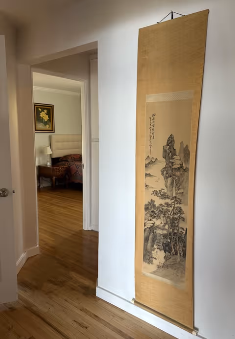 A hallway with wooden flooring and white walls featuring a vertical hanging scroll with traditional Asian landscape art. Through an open doorway, a bedroom is visible with a bed, a nightstand, a lamp, and a framed floral painting on the wall.