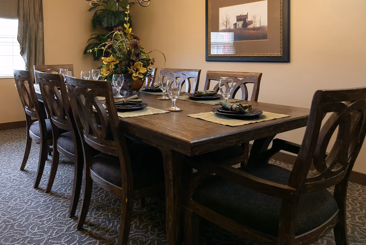 A dining room with a long wooden table set for six people. Each place setting includes a plate, a folded green napkin, and a clear water glass. There is a large floral centerpiece on the table. The room has beige walls, a patterned carpet, a window with curtains, and a framed picture on the wall.