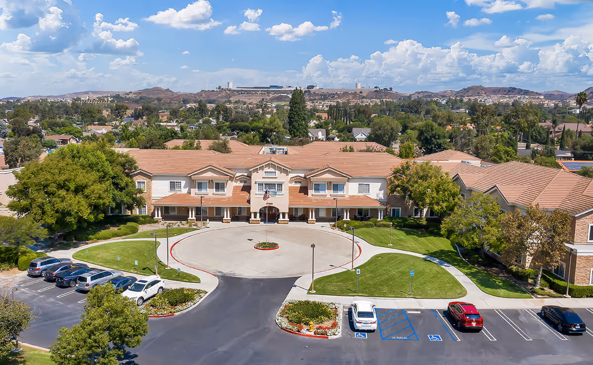 Aerial view of a senior living facility named Bayshire Yorba Linda, showing a large building with a circular driveway and surrounding parking lot. The building has a tan roof and beige exterior with multiple windows. There are landscaped green lawns, trees, and a backdrop of hills and a partly cloudy sky.