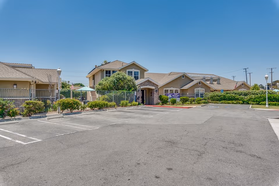 Exterior view of Newport Mesa Senior Living facility showing a large parking lot in the foreground, landscaped bushes, and a beige building with multiple rooflines under a clear blue sky.