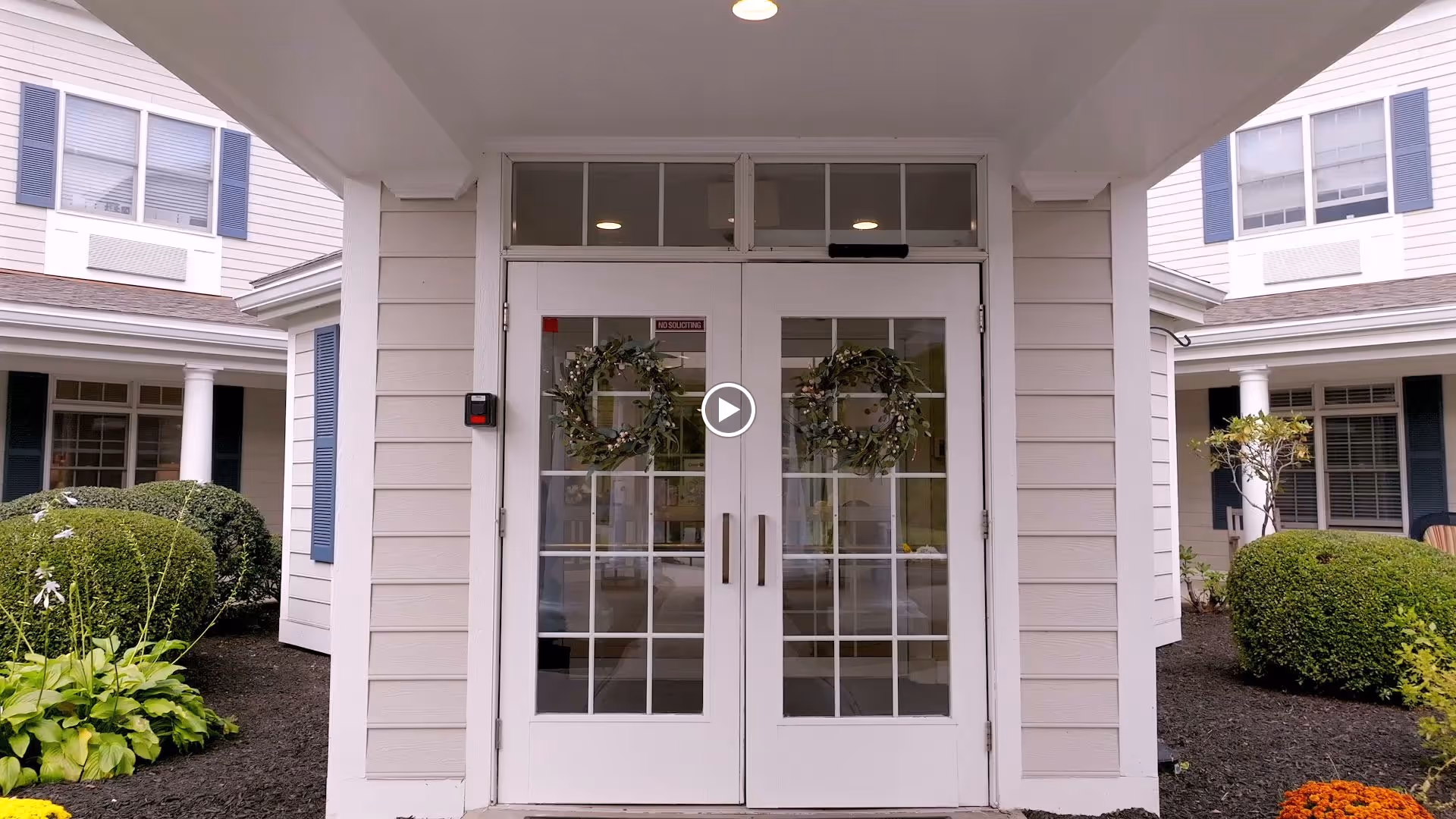 Covered main entrance of the facility with double glass doors adorned with wreaths and flanked by siding and landscaped shrubs.