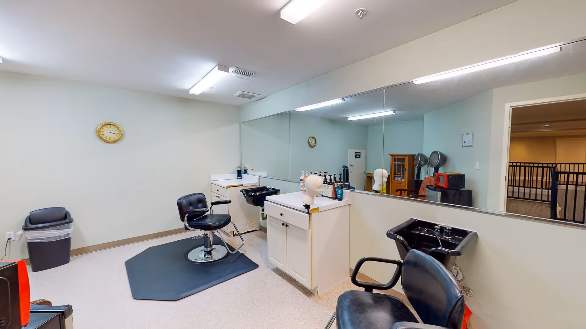 Interior view of a hair salon area in a senior living facility with two black salon chairs, a large mirror on the wall, a white cabinet with hair care products, a black hair washing sink, and a clock on the wall.
