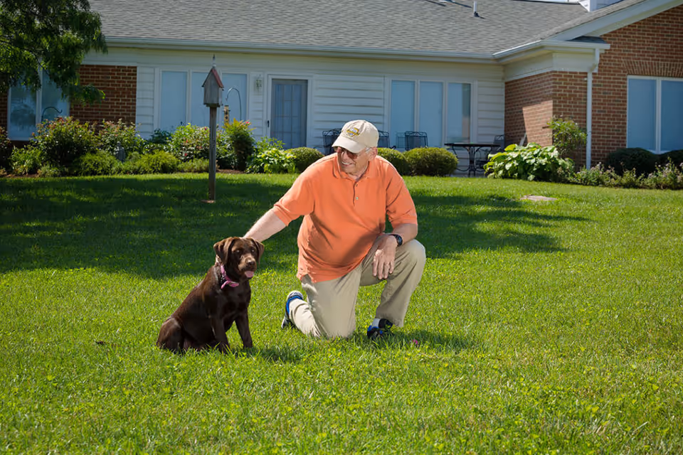 An elderly man wearing an orange shirt, beige pants, and a beige cap kneels on a green lawn outside a building, petting a brown dog sitting beside him. The building has white siding and brick accents with windows and a door visible in the background.