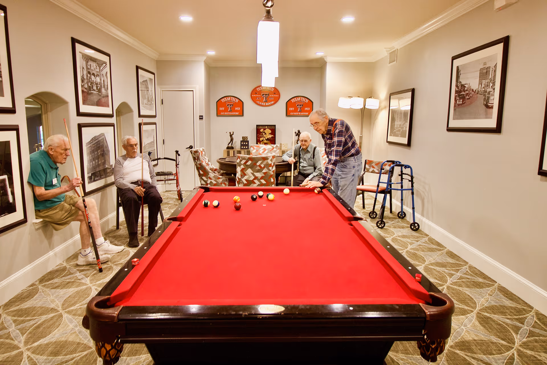 Four elderly men in a game room with a red pool table. One man is playing pool while the others watch. The room has framed black and white photos on the walls, patterned carpet, and comfortable chairs. There are Texas Tech University signs on the back wall and a walker near one of the men.