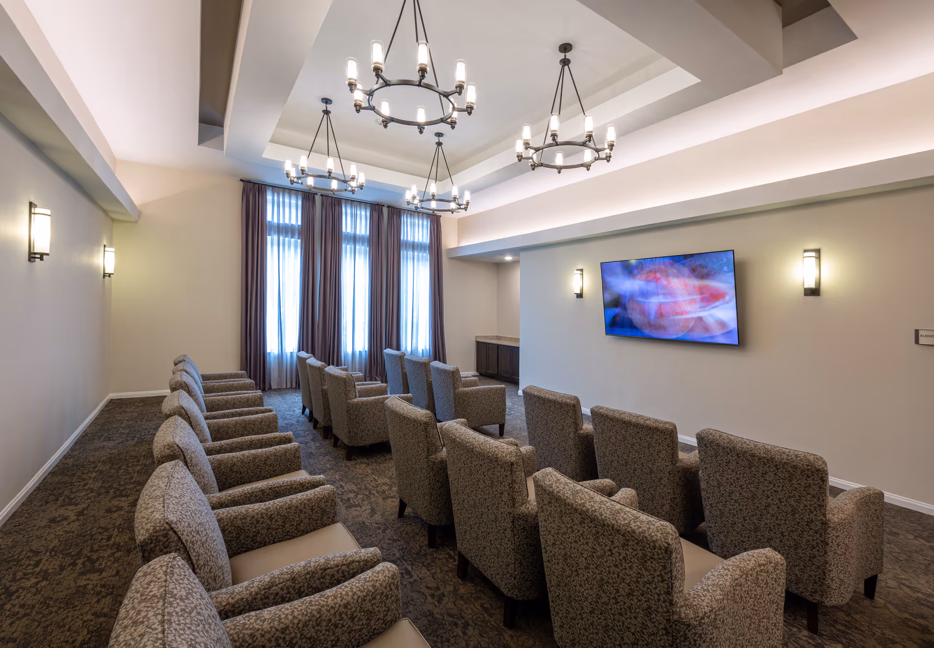 A well-lit room with two rows of comfortable upholstered armchairs facing a wall-mounted flat screen TV. The room has large windows with sheer curtains, three elegant chandeliers hanging from a tray ceiling, and wall sconces providing additional lighting. The carpeted floor and neutral wall colors create a cozy atmosphere.