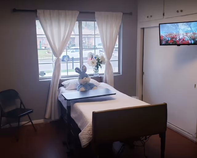 A single bed in a bright bedroom with white curtains at the window, a blue stuffed bunny on the bed, a wall-mounted TV and a folding chair.