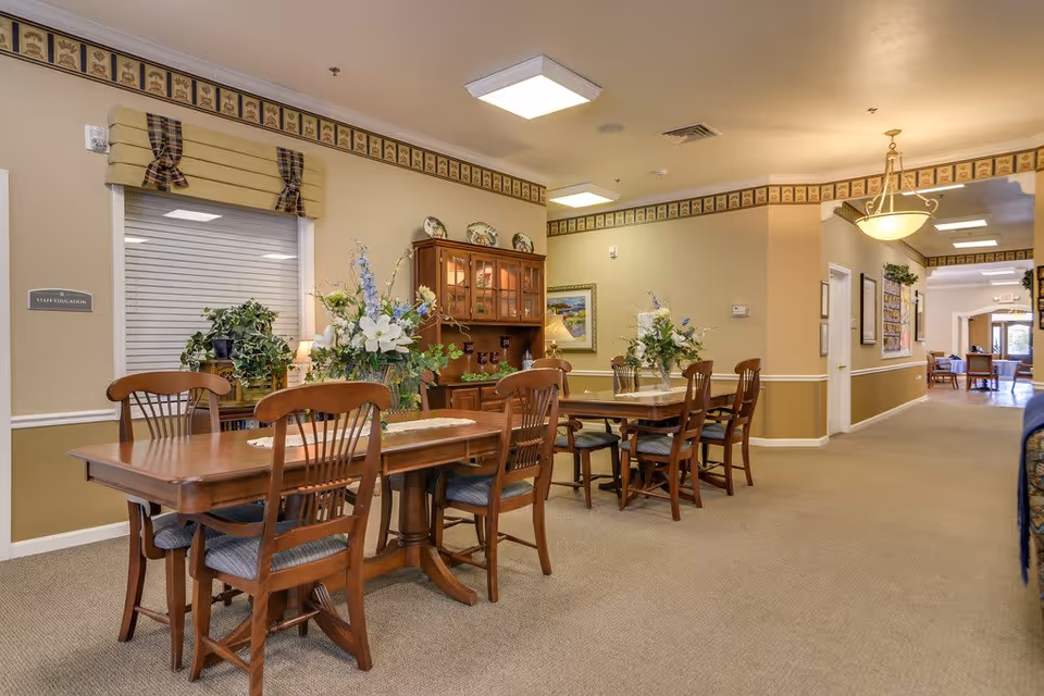 Interior view of a senior living facility dining area with wooden tables and chairs arranged neatly. The tables are decorated with floral centerpieces. There is a wooden cabinet with decorative plates and glassware against the wall. The walls have a beige and light brown color scheme with a decorative border near the ceiling. A hallway extends to the right with additional seating visible in the distance.