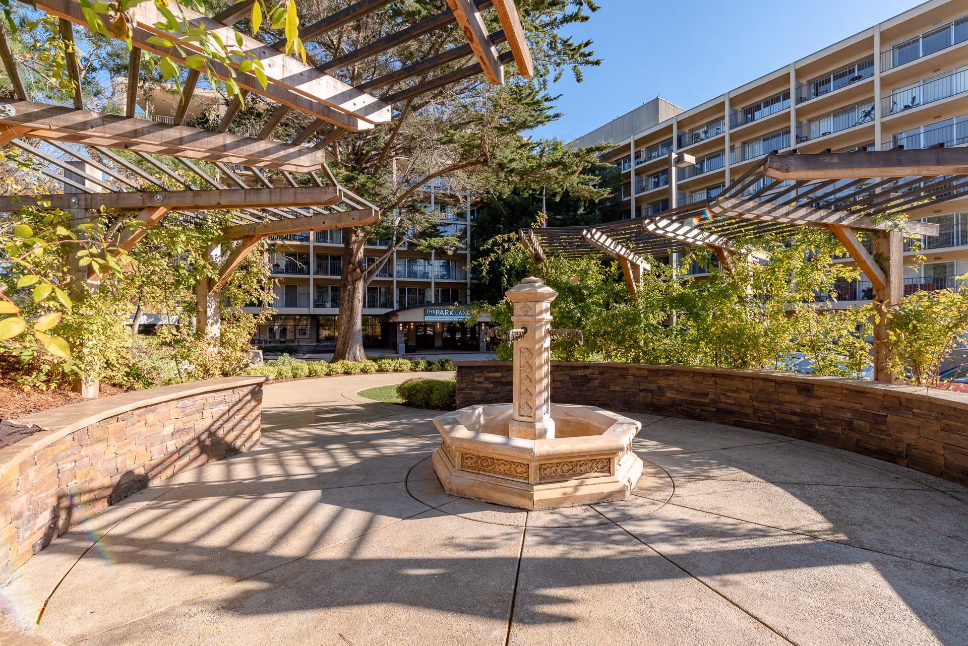 Outdoor courtyard area at The Park Lane senior living facility featuring a stone water fountain in the center, surrounded by curved stone benches and wooden pergolas with climbing plants. The multi-story building with balconies is visible in the background under a clear blue sky.
