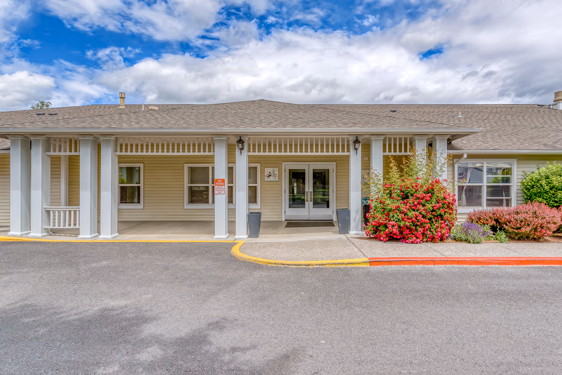 Front entrance of Carriage Place Memory Care facility with beige siding, white columns, double glass doors, and a porch area. There are red and green bushes to the right of the entrance and a cloudy blue sky above.
