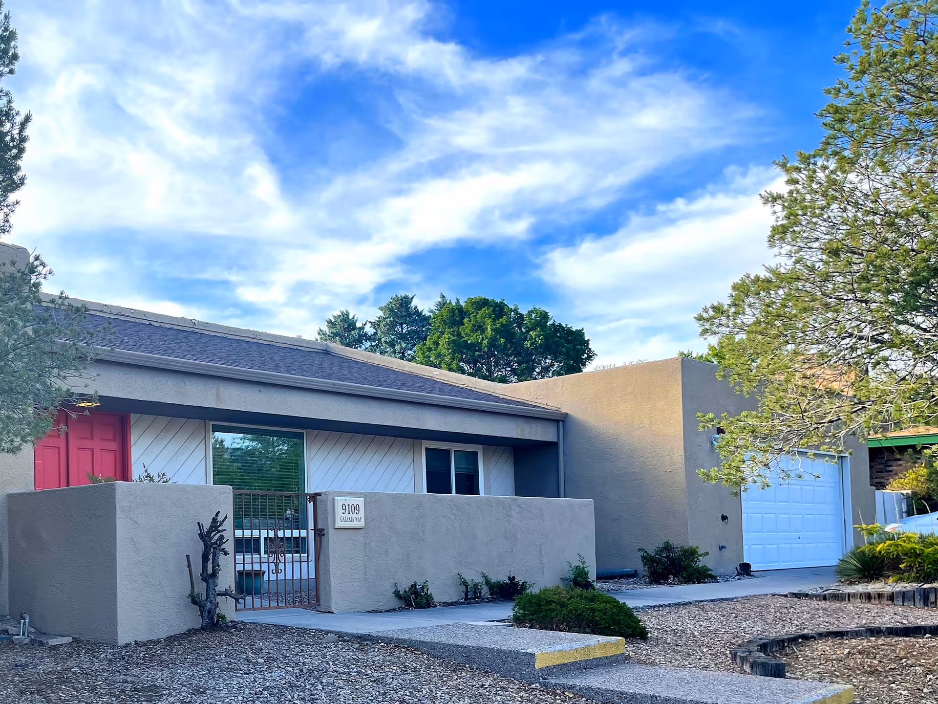Single-story stucco house with a gated front patio, red door, and attached garage under a blue sky.