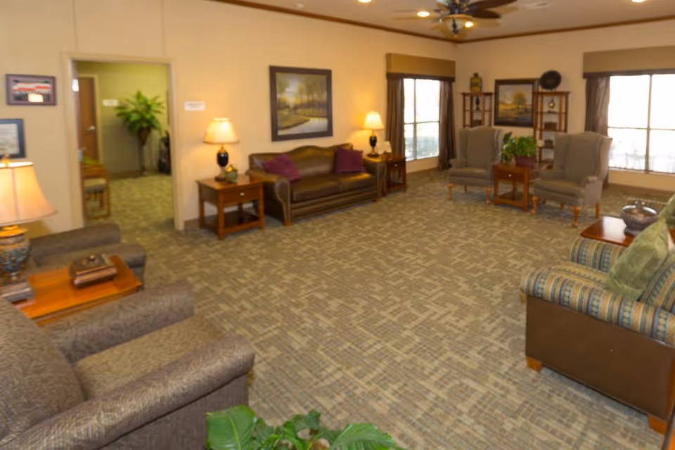 A spacious living room area in a senior living facility with multiple armchairs, a sofa, side tables with lamps, framed artwork on the walls, large windows with curtains, and carpeted flooring. There is a ceiling fan with lights and some indoor plants adding greenery to the space.