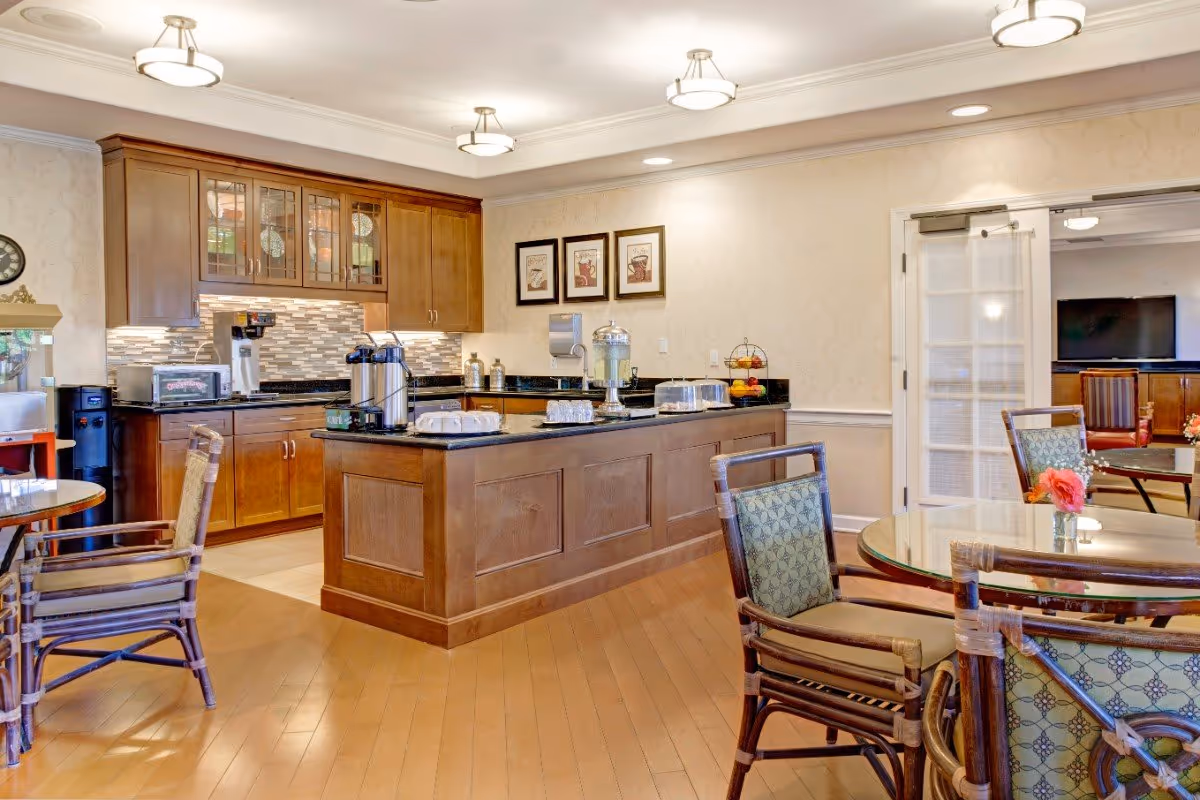 Well-lit communal dining area with a central serving island, wooden cabinetry, and round glass-top tables with chairs.