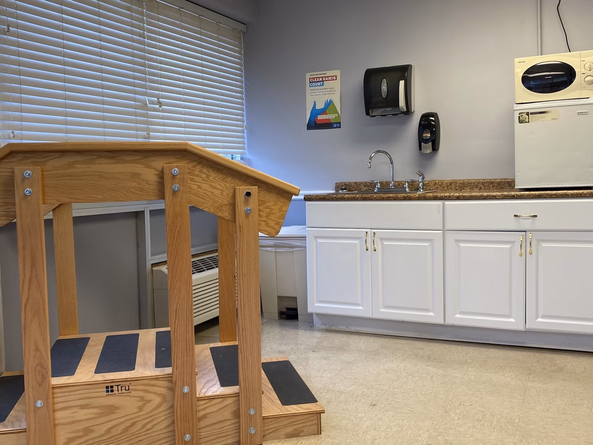 A room with a wooden handrail and steps in the foreground, a window with closed blinds, a countertop with a sink, white cabinets, a microwave, a paper towel dispenser, and a soap dispenser mounted on the wall. A poster about clean hands is also visible on the wall.