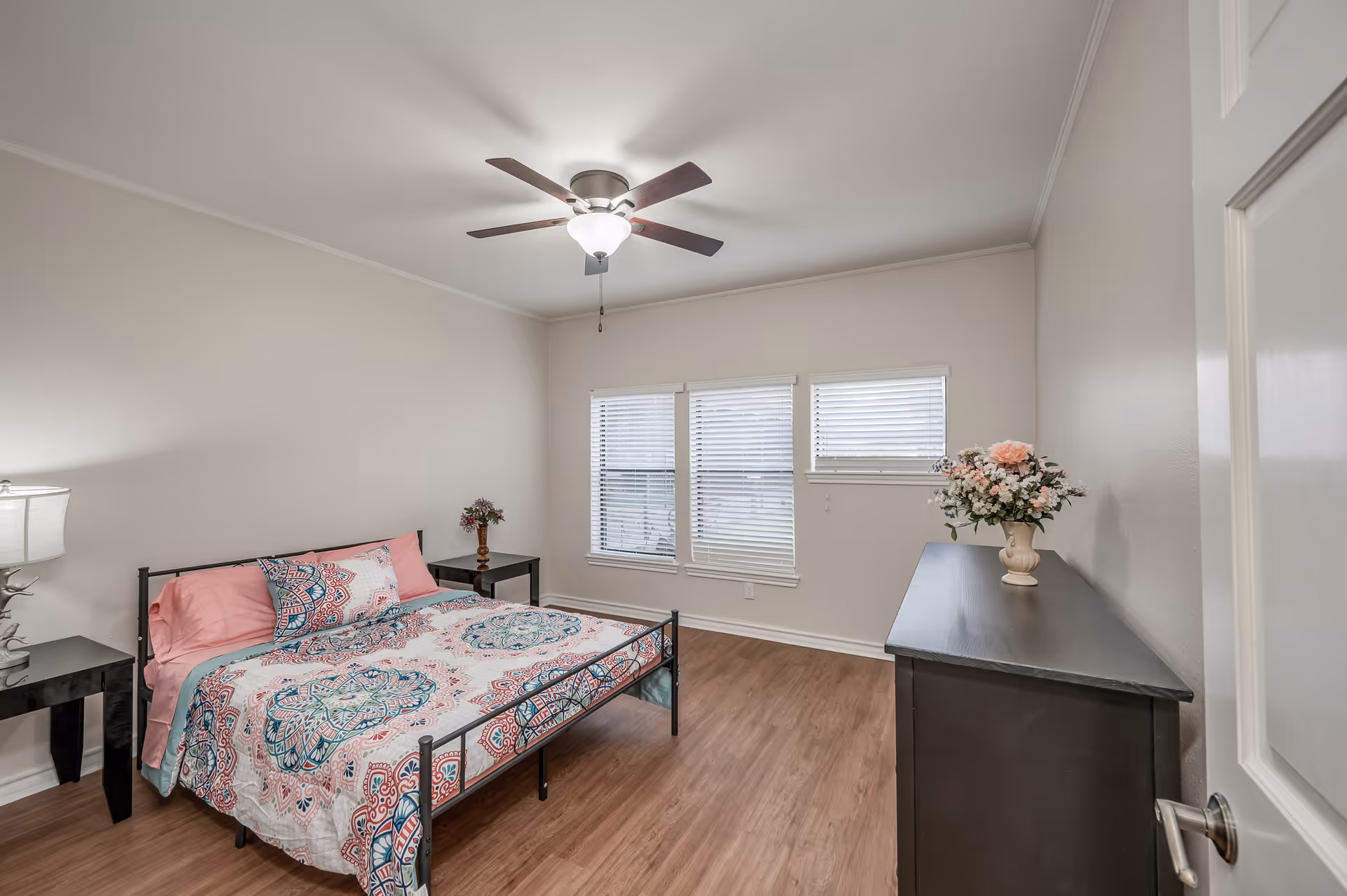 A bright bedroom with a bed featuring a colorful patterned bedspread and pink pillows, two black nightstands with lamps and flowers, a ceiling fan with light, three windows with blinds, and a black dresser with a vase of flowers on top.
