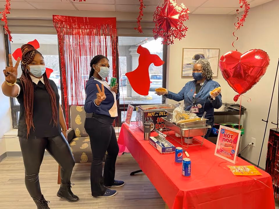 Three women wearing face masks stand around a table decorated with red Valentine-themed decorations including a heart-shaped balloon and red hanging ornaments. Two women on the left are making peace signs, while the woman on the right is holding two hot dog buns behind a table with a hot dog warmer, soda cans, and other food items. The setting appears to be an indoor common area with large windows in the background.