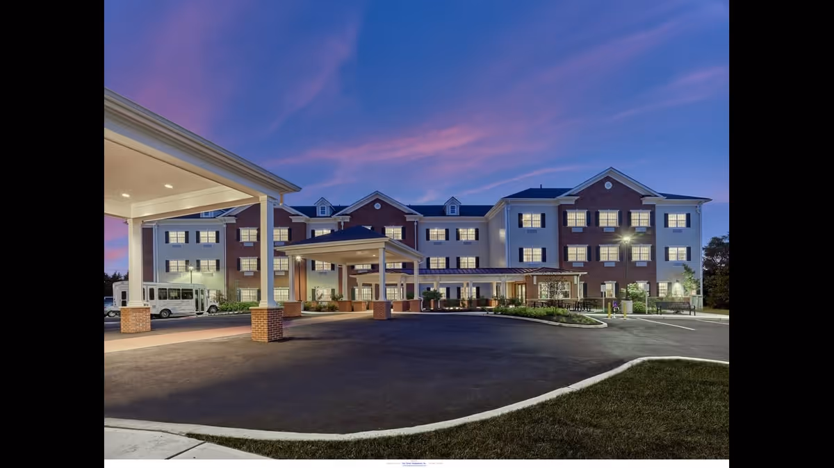 Exterior view of The Chelsea at Fair Lawn senior living facility at dusk, showing a large multi-story building with illuminated windows, covered entrance with pillars, a driveway, and a shuttle bus parked on the left side.