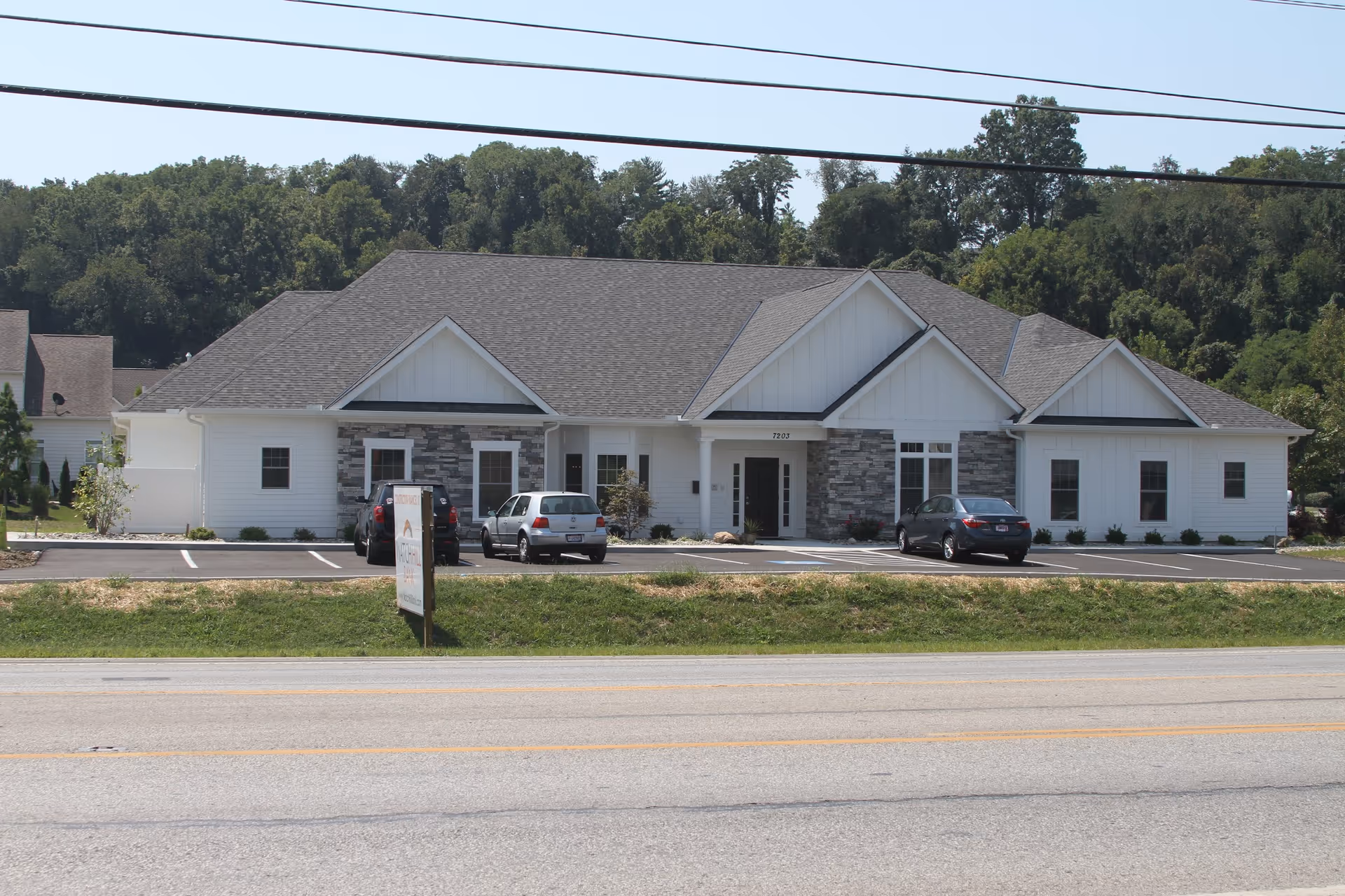 Front exterior of a single-story white assisted living building with stone accents, a parking lot with cars, and trees behind it.