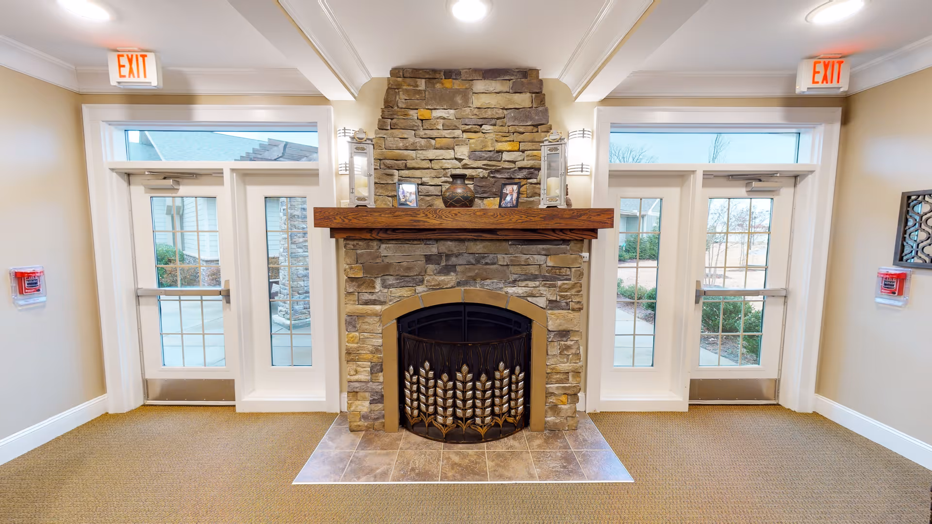 Interior view of a common area in Oakview Park featuring a stone fireplace with a wooden mantel. On the mantel are two lanterns, a decorative vase, and two framed photos. There are two glass doors on either side of the fireplace leading outside, each with an exit sign above. The walls are painted beige, and the floor is carpeted.