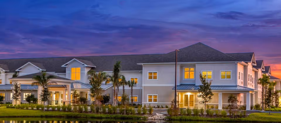 Two-story residential building at dusk with lit windows, palm trees, landscaped grounds and a reflecting pond in front.