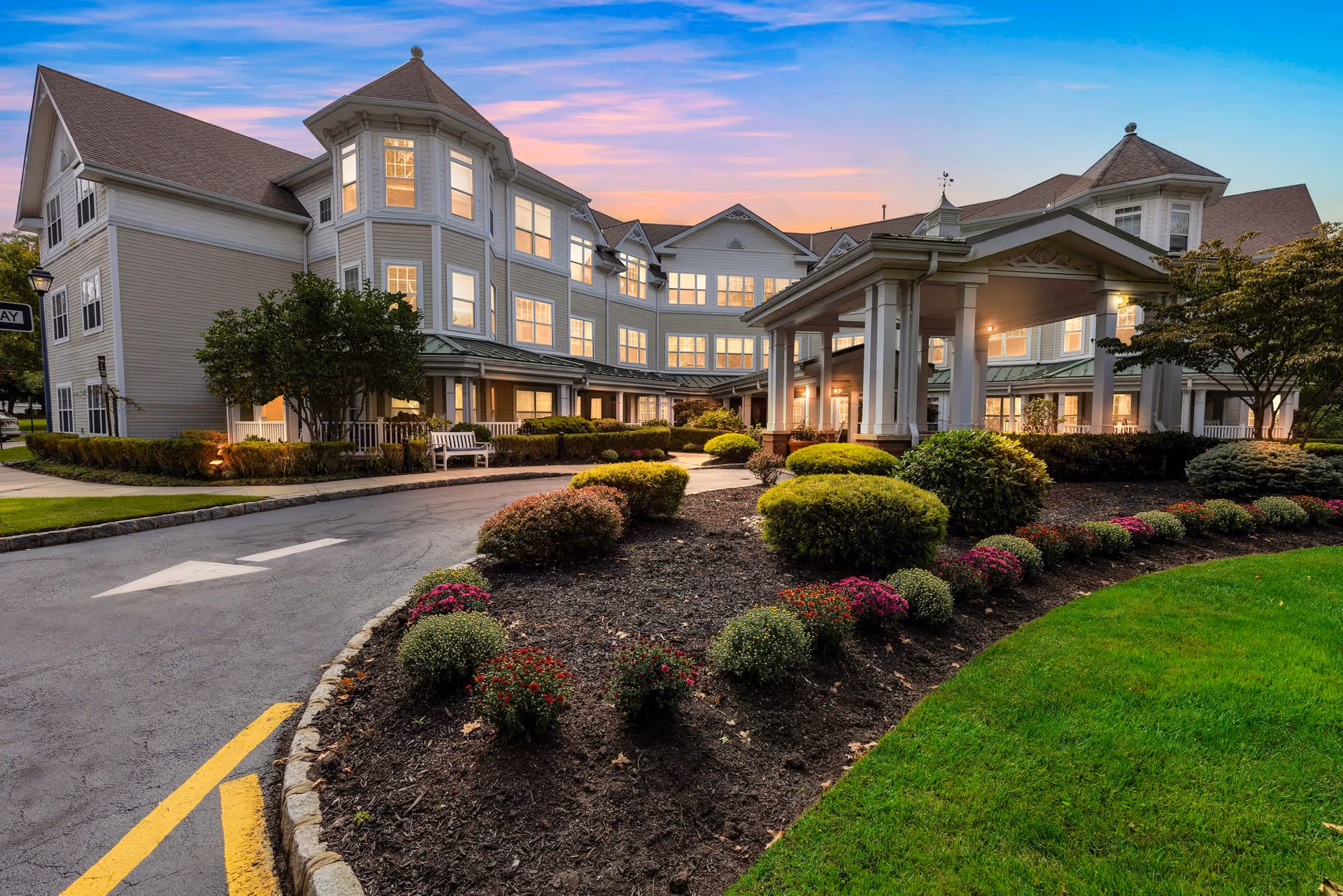 Exterior view of a large, multi-story senior living facility building at sunset with well-maintained landscaping including bushes and flowers, a driveway with directional arrows, and a covered entrance area.
