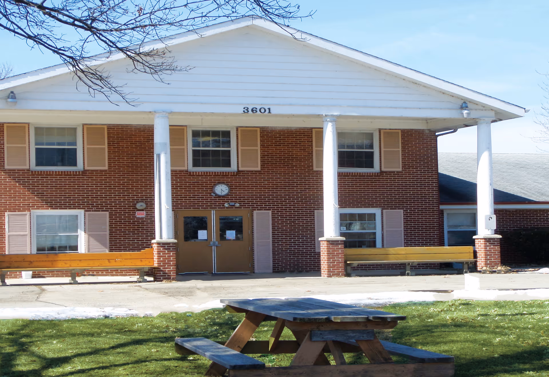 Front exterior view of a two-story brick building with white columns and a white triangular pediment. The building has several windows with beige shutters and two brown double doors at the entrance. There is a clock above the doors and two wooden benches on either side of the entrance. In the foreground, there is a wooden picnic table on a grassy area with some patches of snow.