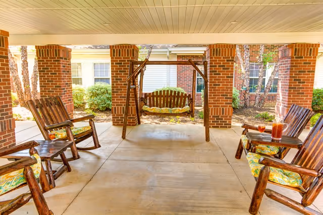 Covered outdoor seating area with wooden rocking chairs and a wooden swing bench, all with patterned cushions. The area is supported by brick columns and overlooks a garden with bushes and trees.