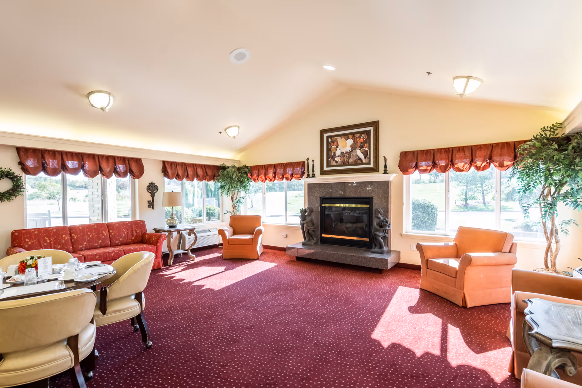 A bright and spacious senior living common area with large windows letting in natural light. The room features a red carpet, a red sofa, two orange armchairs, a round dining table with cream-colored chairs, a fireplace with decorative statues on either side, and framed artwork above the fireplace. There are also plants and red valance curtains on the windows.