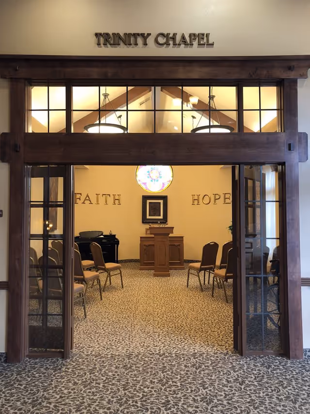 Entrance to the Trinity Chapel with wooden framed doors opening to a small chapel containing chairs, a pulpit, a stained-glass window and the words "FAITH" and "HOPE" on the back wall.