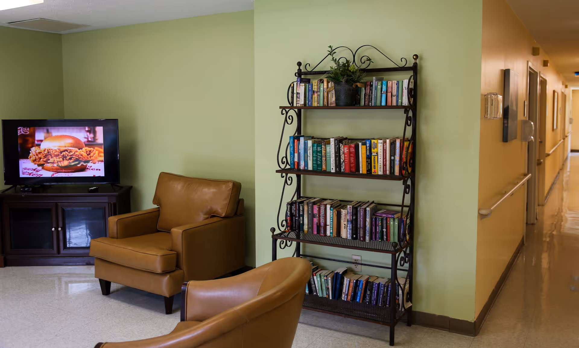 A cozy seating area in a senior living facility with two brown leather armchairs, a black metal bookshelf filled with books, and a television on a dark wooden stand displaying an image of a sandwich. The walls are painted light green and there is a hallway visible to the right.