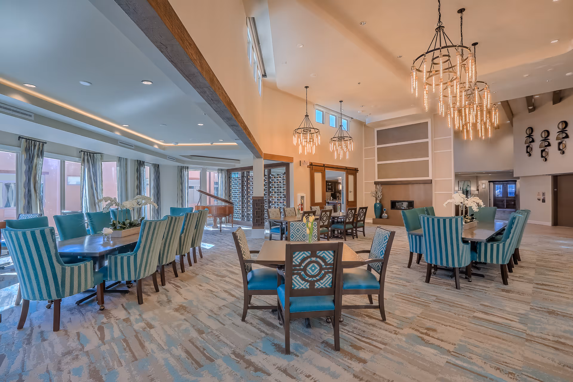Spacious, well-lit communal dining and lounge area with teal upholstered chairs, multiple tables, chandeliers, and a piano.