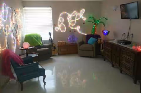 Common living room in a senior facility with neon art, chairs, a TV, and a dresser.