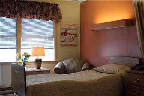 Senior living bedroom with single bed, chair, nightstand, lamp, and window blinds