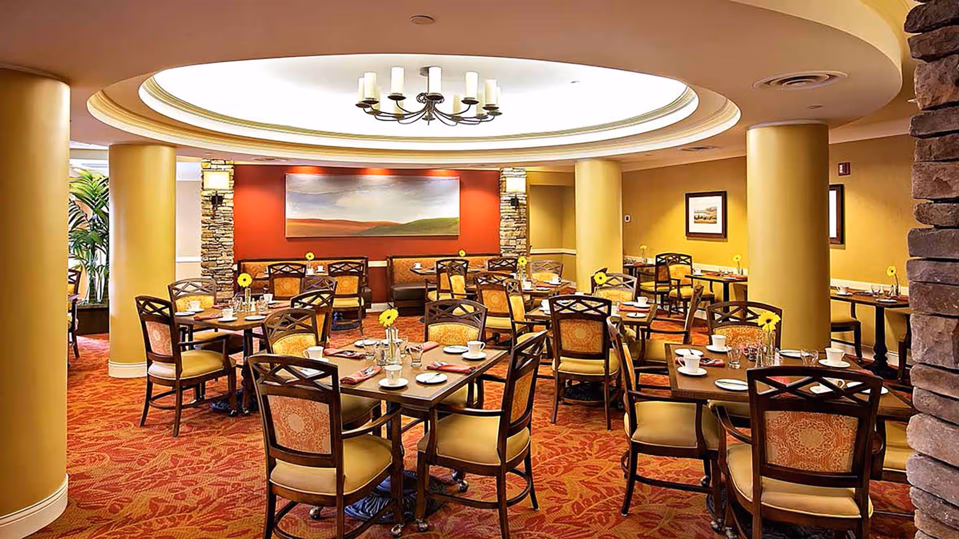 Warm, warmly lit dining room with multiple set tables and chairs, yellow columns, and a central circular ceiling with chandelier.