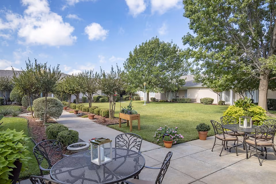 Sunny outdoor courtyard with metal patio tables and chairs, potted plants, a walkway and lawn surrounded by a single-story building.