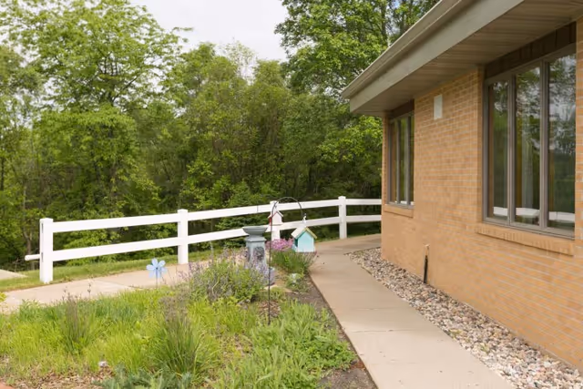 A paved walkway beside a brick building with windows, bordered by a garden with green plants and a white wooden fence. Trees and greenery are visible in the background under a cloudy sky.
