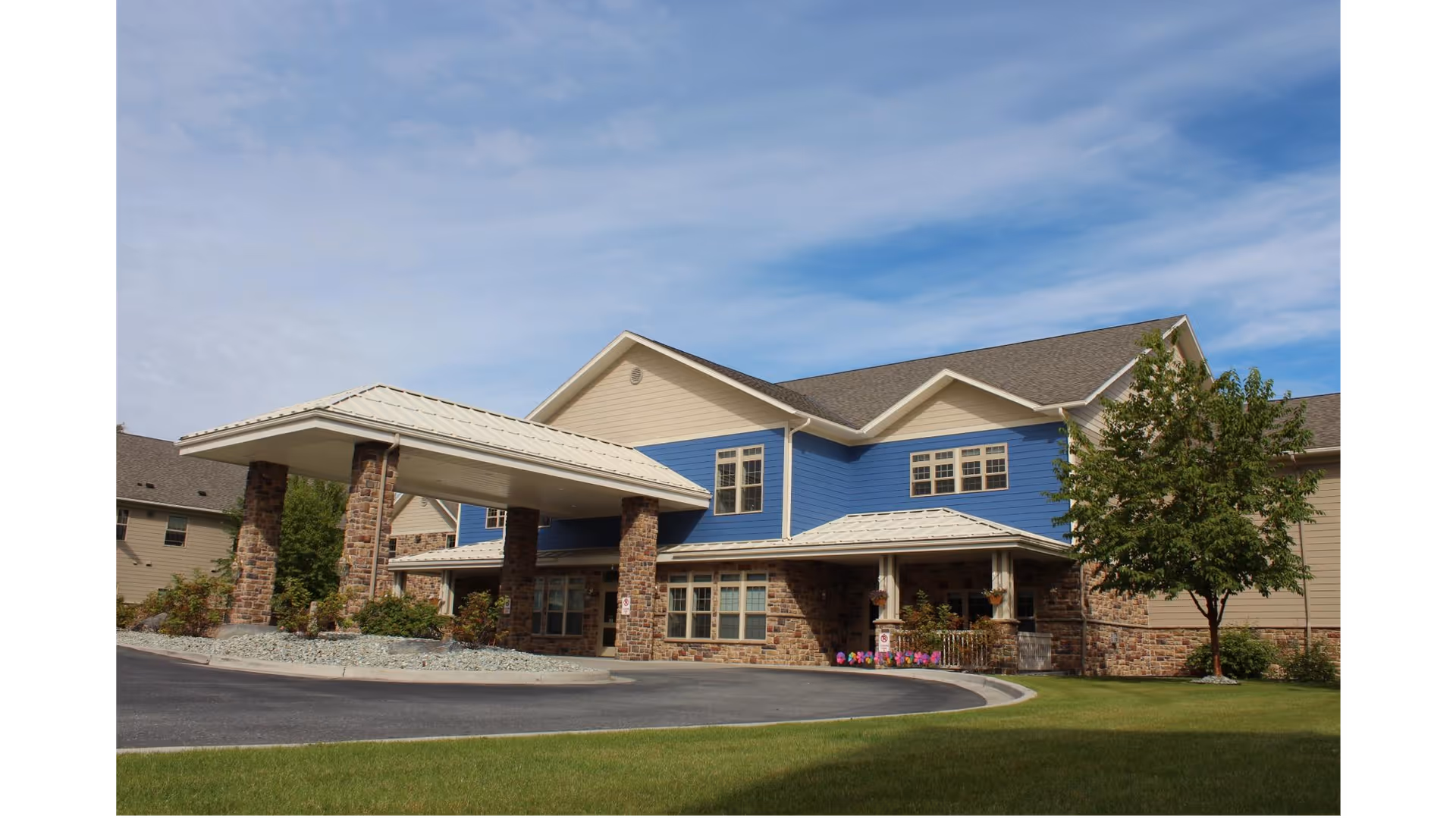 Front exterior of a two-story retirement community building with a covered porte-cochere, stone columns, blue siding and a circular driveway.