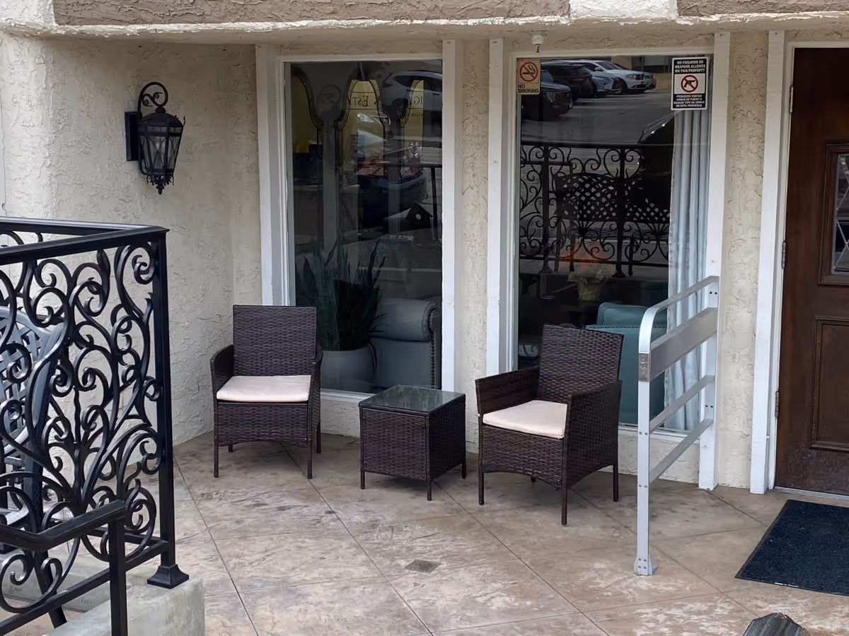 Outdoor patio area with two wicker chairs with cushions and a small glass-top table between them. The patio has a textured beige wall, a decorative black metal railing on the left, a wall-mounted lantern light fixture, two large windows with reflections of parked cars, and a wooden door on the right with a black doormat. There is also a metal safety railing near the door.