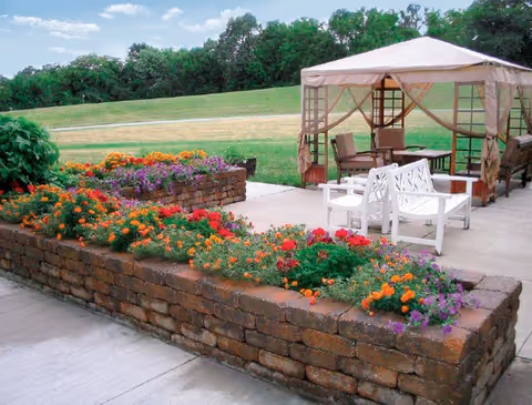 Outdoor patio area with a brick raised flower bed filled with colorful flowers including orange, red, and purple blooms. There is a beige canopy gazebo with mesh sides and several chairs underneath it. The background shows a grassy hill and trees.