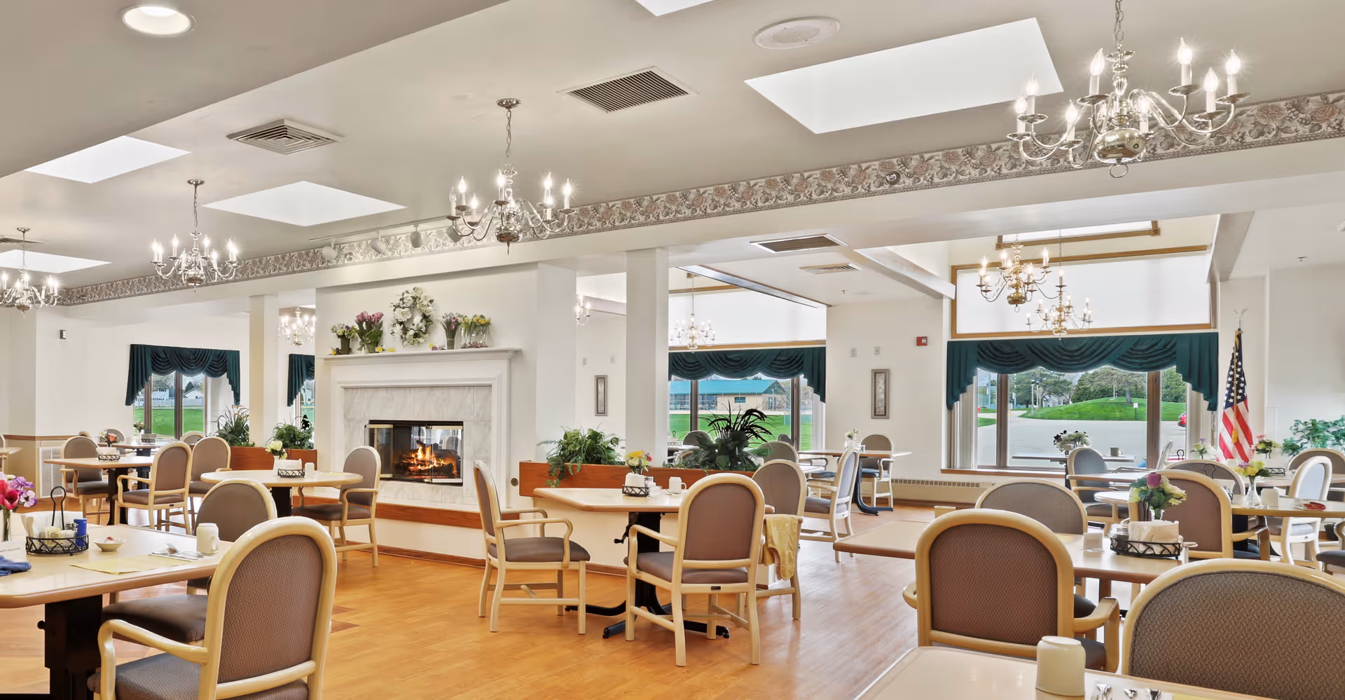 Bright senior living dining room with tables and chairs, chandeliers, a fireplace, and large windows.