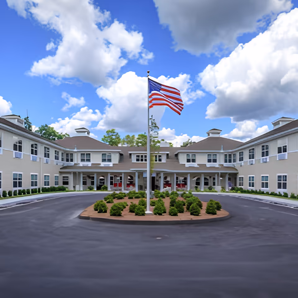 Front entrance of a two-story assisted living building with a circular driveway and an American flag on a central flagpole.