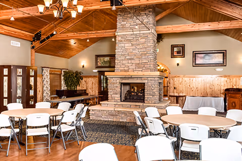 A spacious common area with round tables and white folding chairs arranged around a large stone fireplace. The room features wooden walls and ceiling beams, a chandelier, framed artwork on the walls, and a piano in the corner.