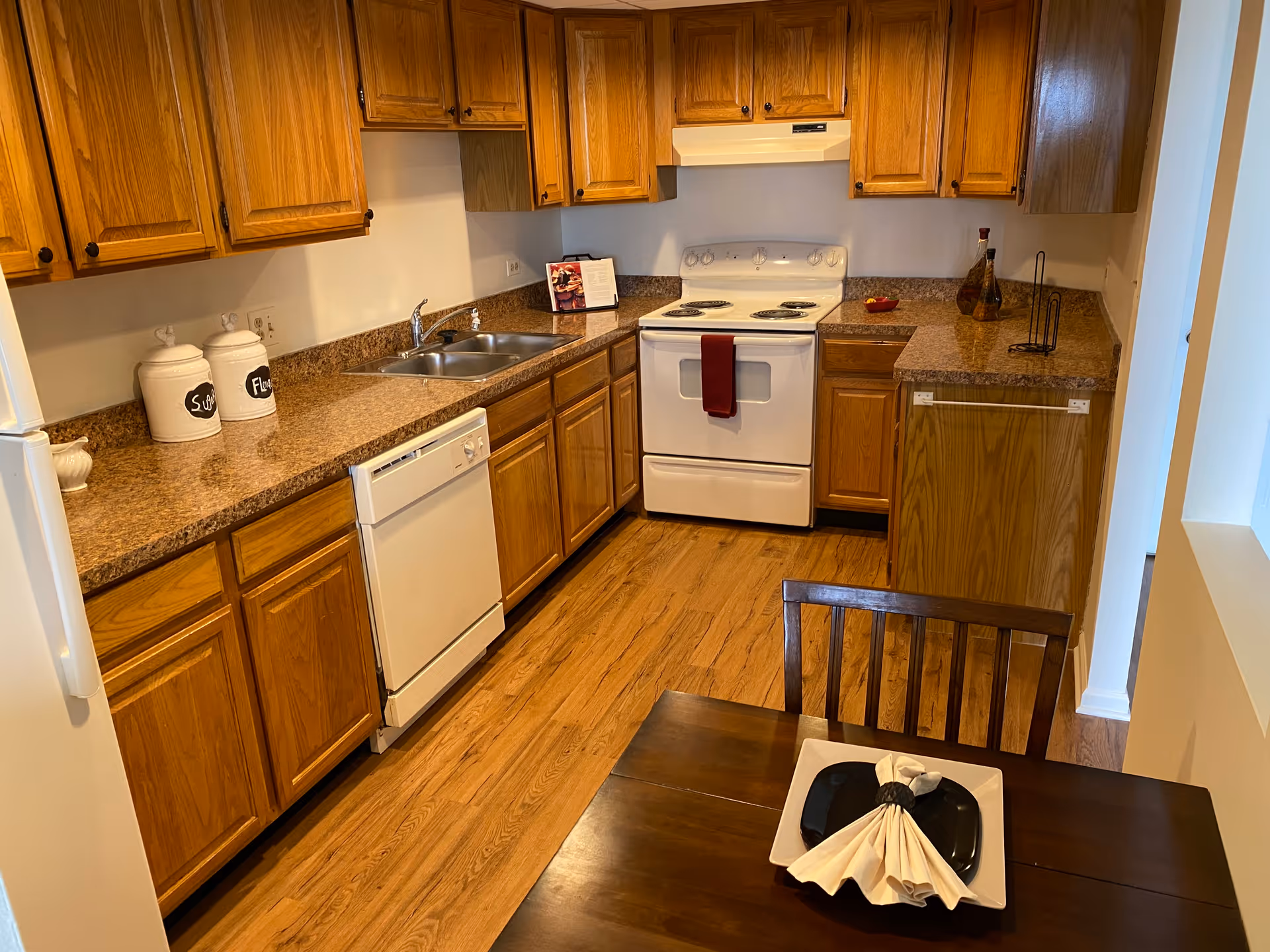 Well-lit kitchen with oak cabinets, laminate countertops, a white stove and dishwasher, double sink, and a small dining table in the foreground.
