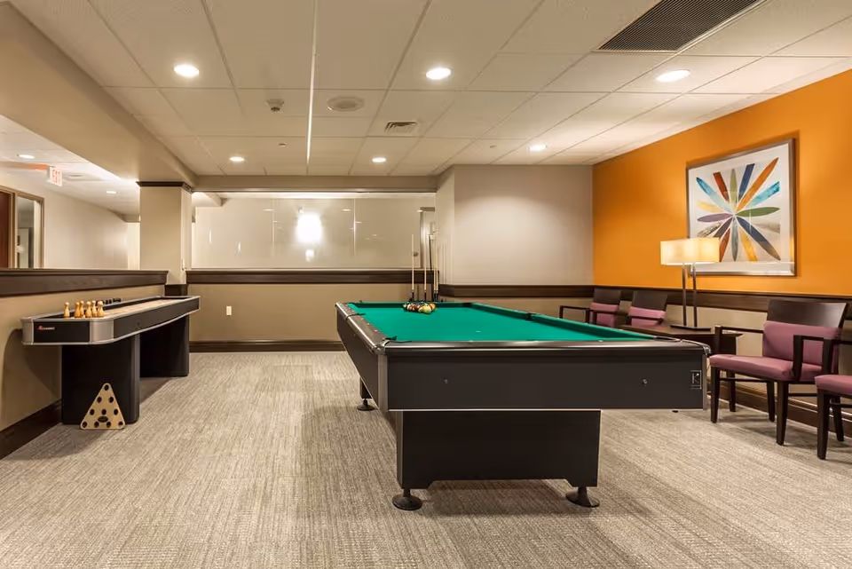 A recreational room with a pool table in the center, a shuffleboard game on the left, and several chairs with purple cushions along the right wall. The walls are painted beige and orange, with a colorful abstract artwork and a floor lamp on the right side.