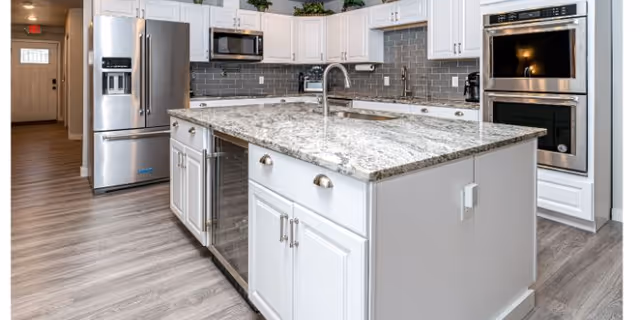 Modern kitchen with a large granite-topped island, white cabinetry, stainless steel appliances, and gray subway tile backsplash.